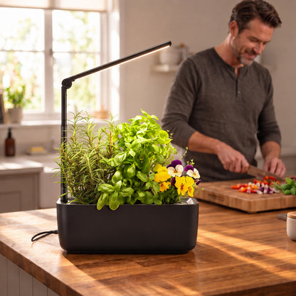 Ermonix LumiGarden 3 in black with black LED grow light arm, shown filled with green herbs and small red/yellow tomatoes on a clean white background, filled with green herbs and small pansies, set on a wooden kitchen island. A man is blurred in the background chopping vegetables in a bright kitchen.